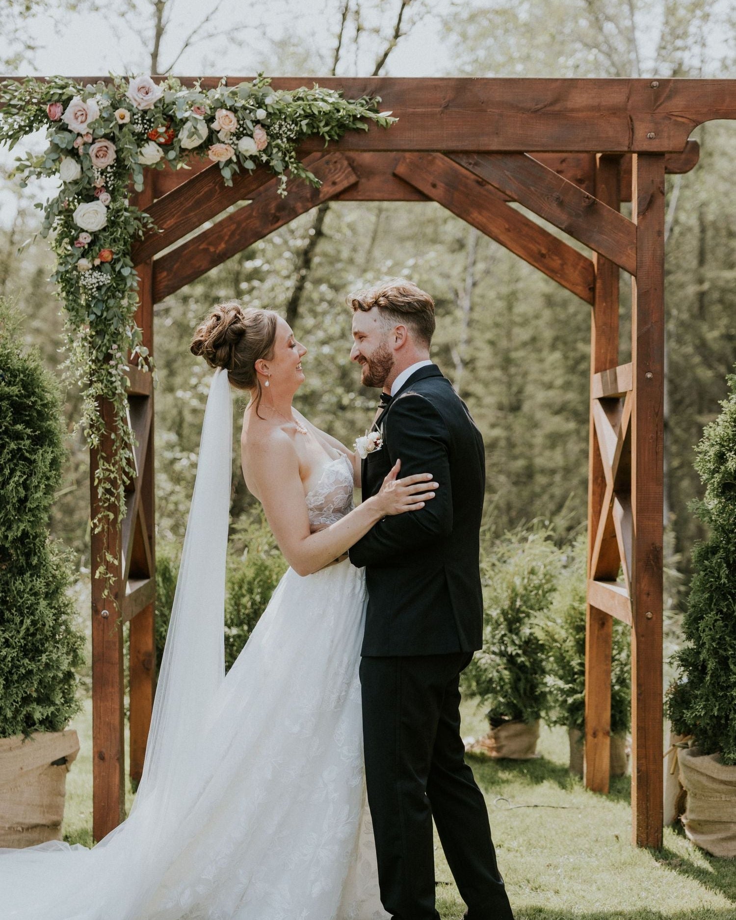 Couple embracing under a wooden arch decorated with flowers, surrounded by greenery.